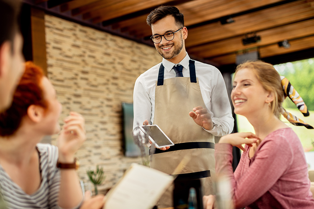 Happy waiter using touchpad while communicating with guests in a restaurant.
